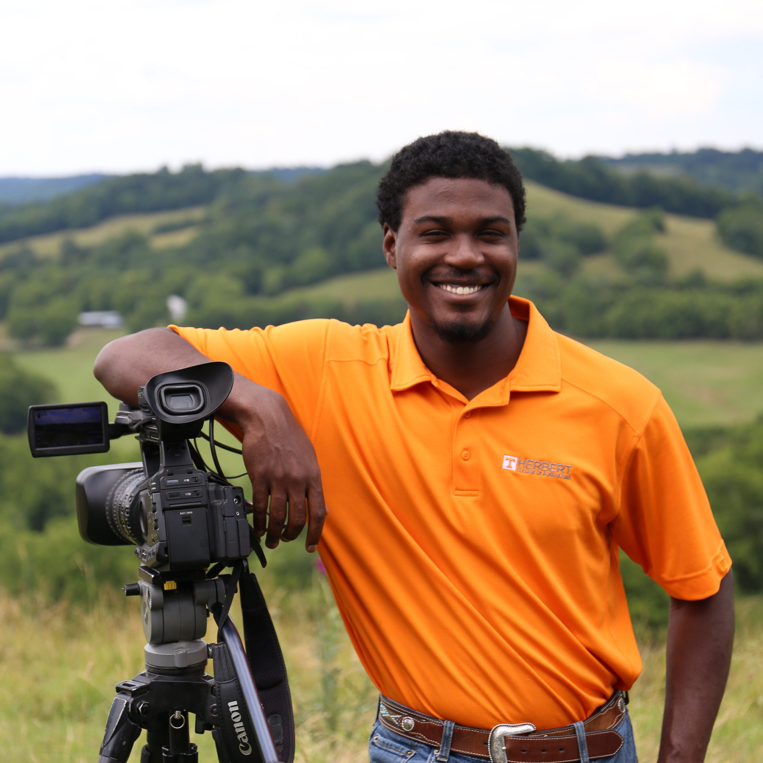 African American male student smiles and leans on a camera mounted on a tripod with the Tennessee hills in the background 